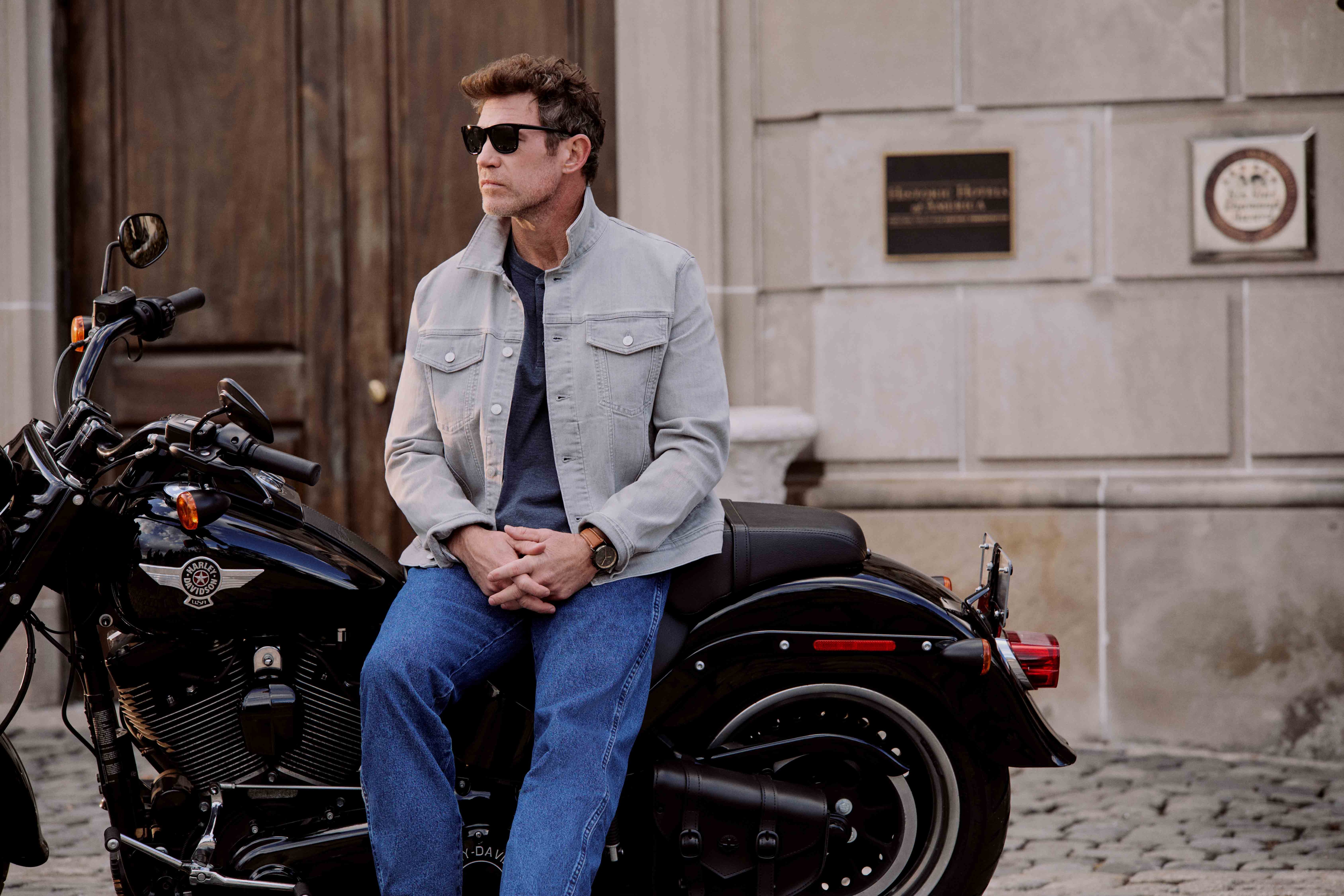 Man in sunglasses and denim jacket sitting on a black motorcycle in front of a stone building