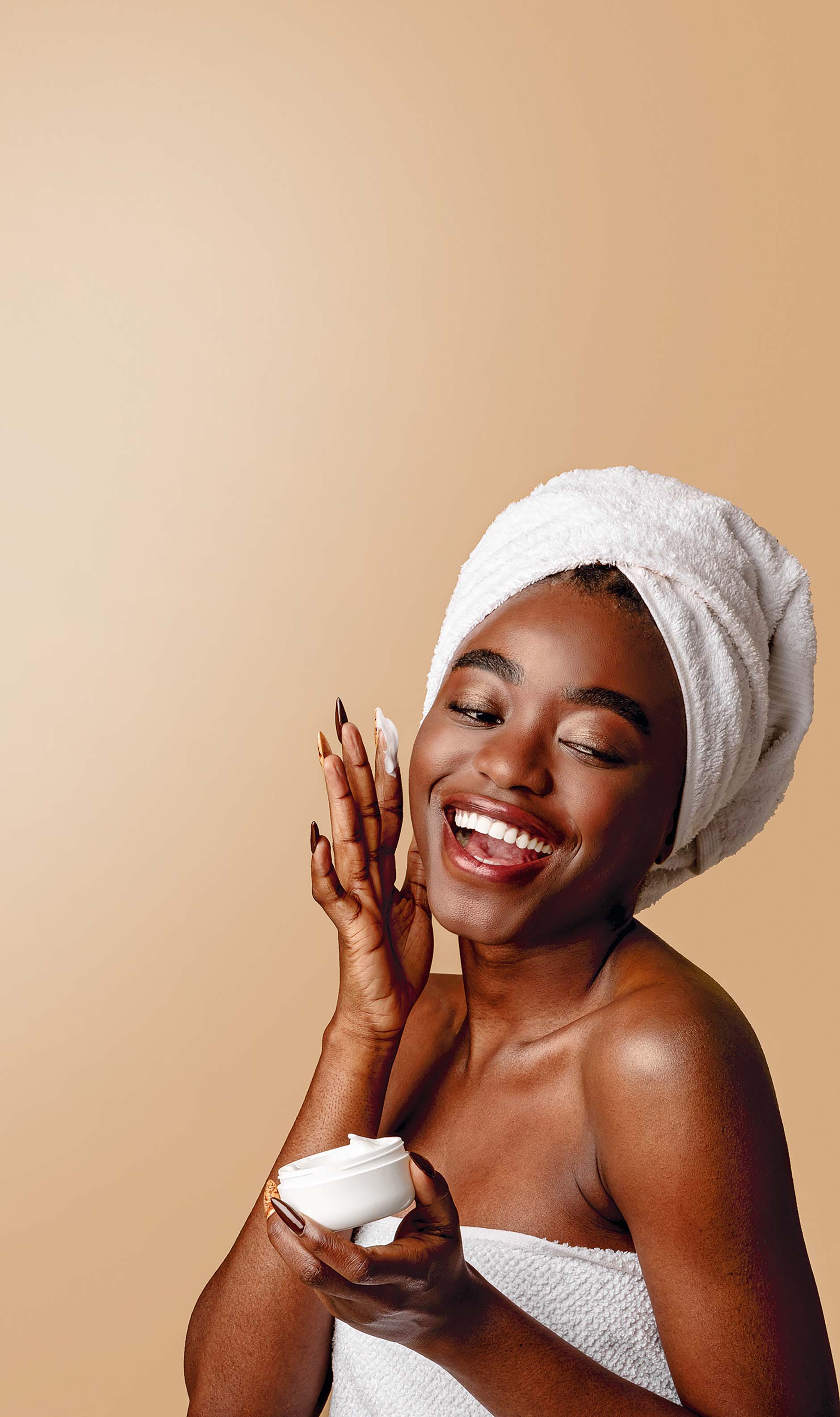 Femme joyeuse en serviette appliquant une crème visage, possiblement une publicité pour produit de soin