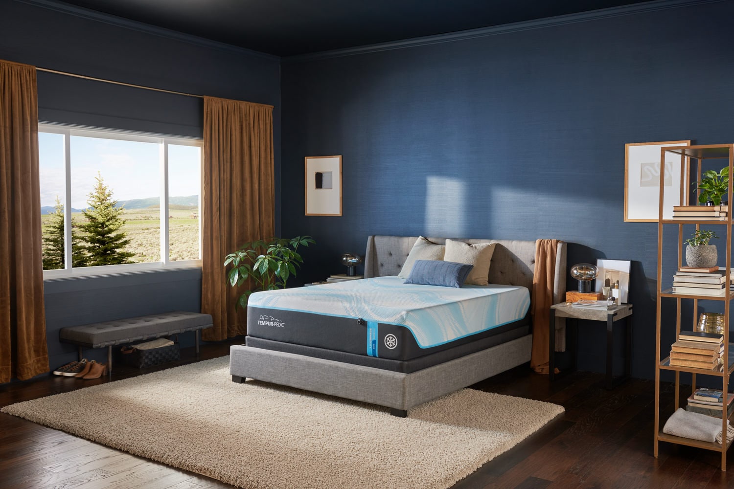 Modern bedroom with a blue theme, featuring a bed, desk, bookshelf, and window view of nature