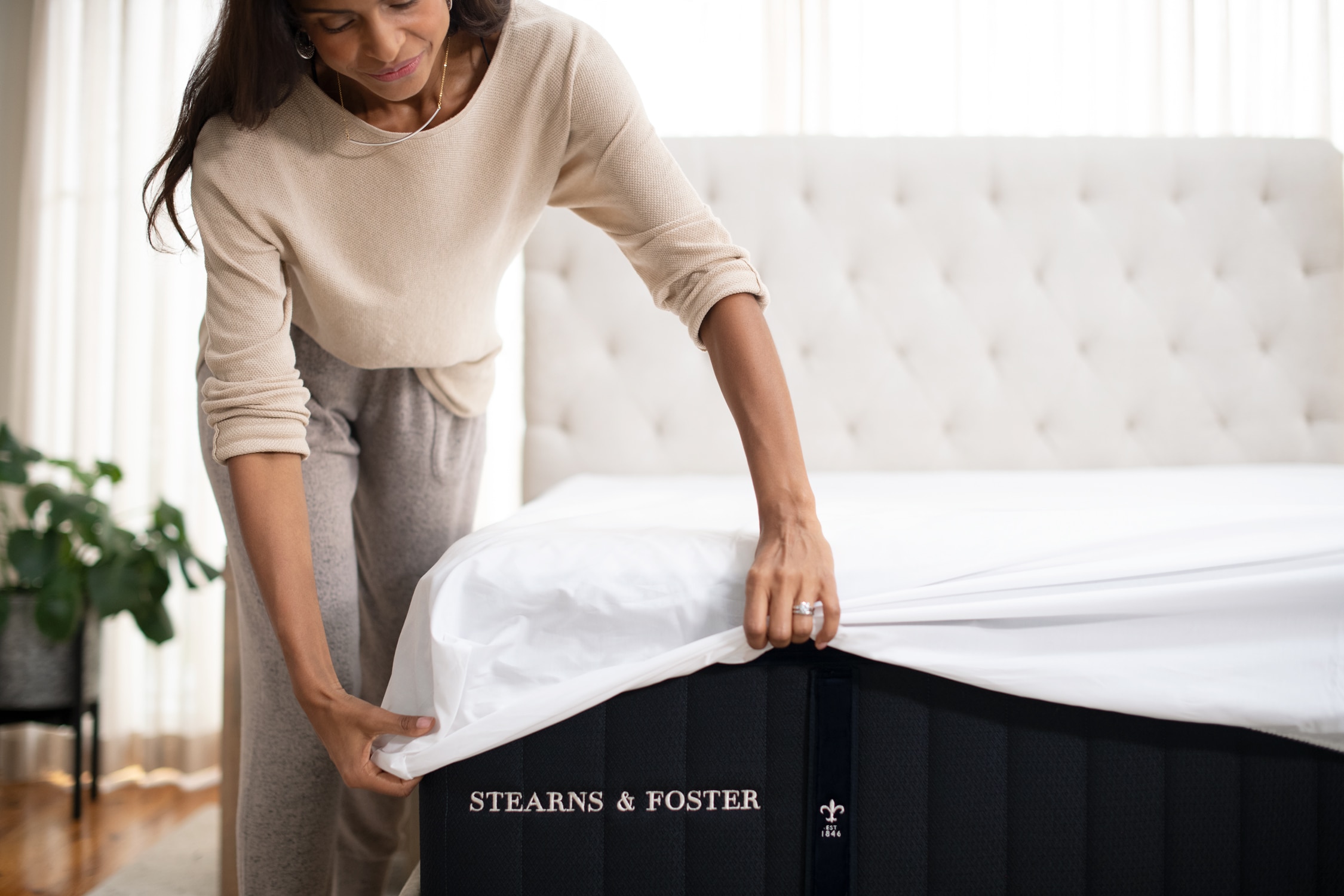 A woman adjusts a white bedsheet on a mattress in a well-lit bedroom