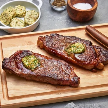 Two grilled steaks with herb butter on a wooden board, spices and salt nearby