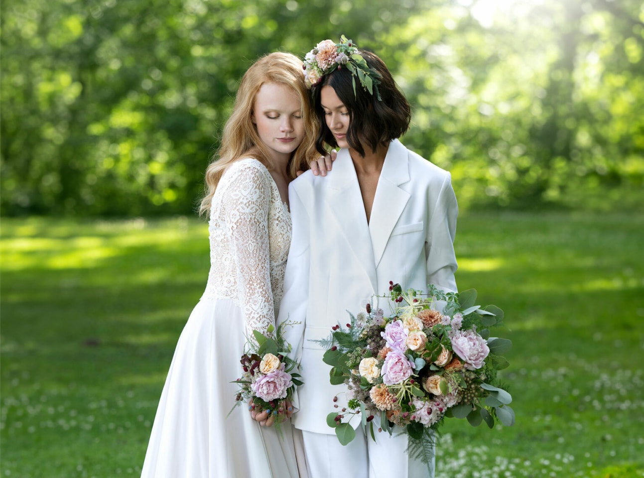 Two women in wedding attire holding bouquets in a sunlit park
