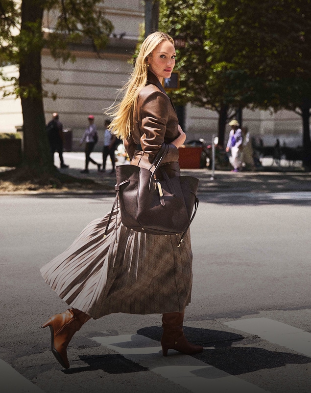 Woman walking across a street wearing a pleated skirt and carrying a large bag