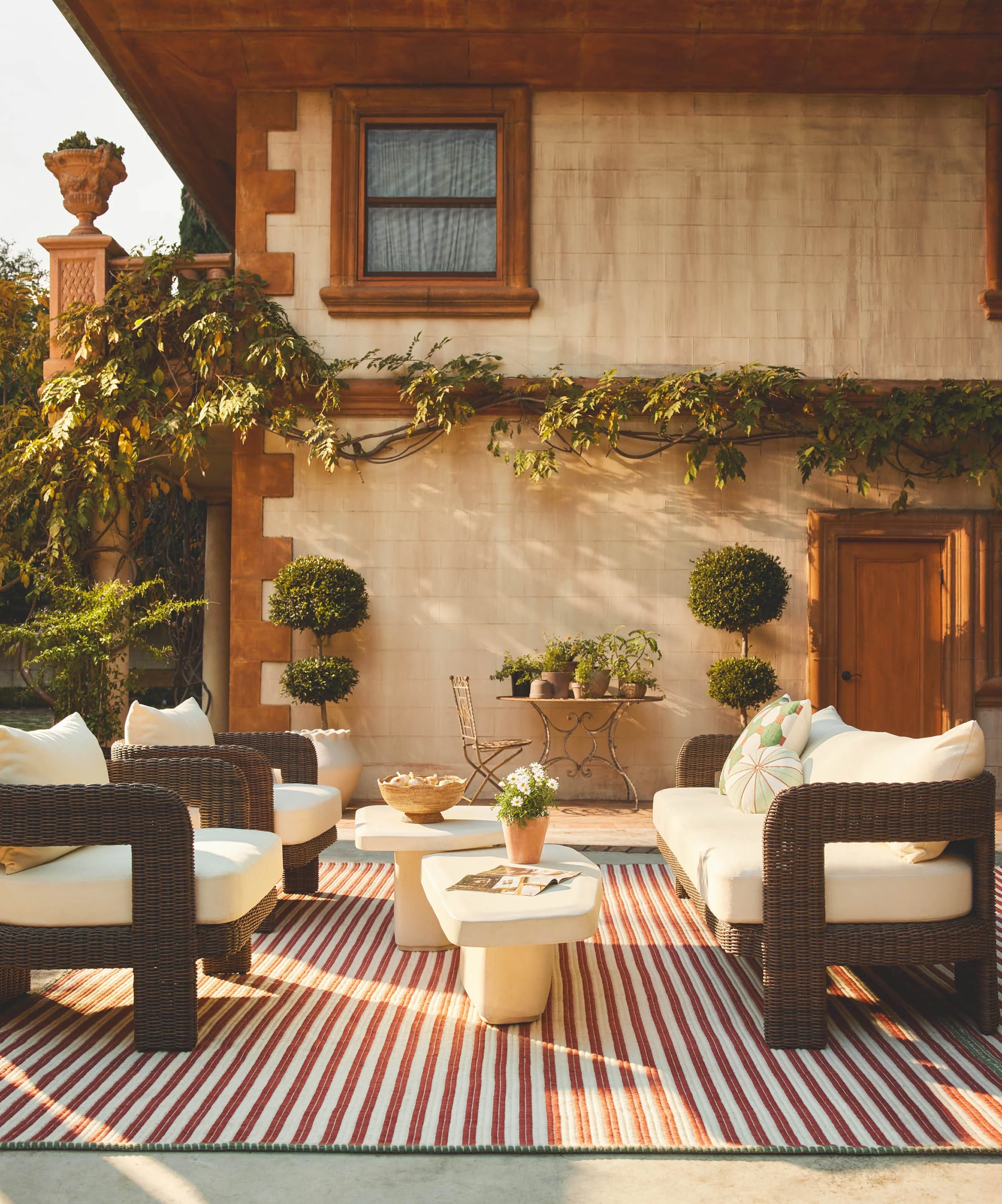 Cozy outdoor patio area with white furniture, striped rug, and greenery against a rustic house facade