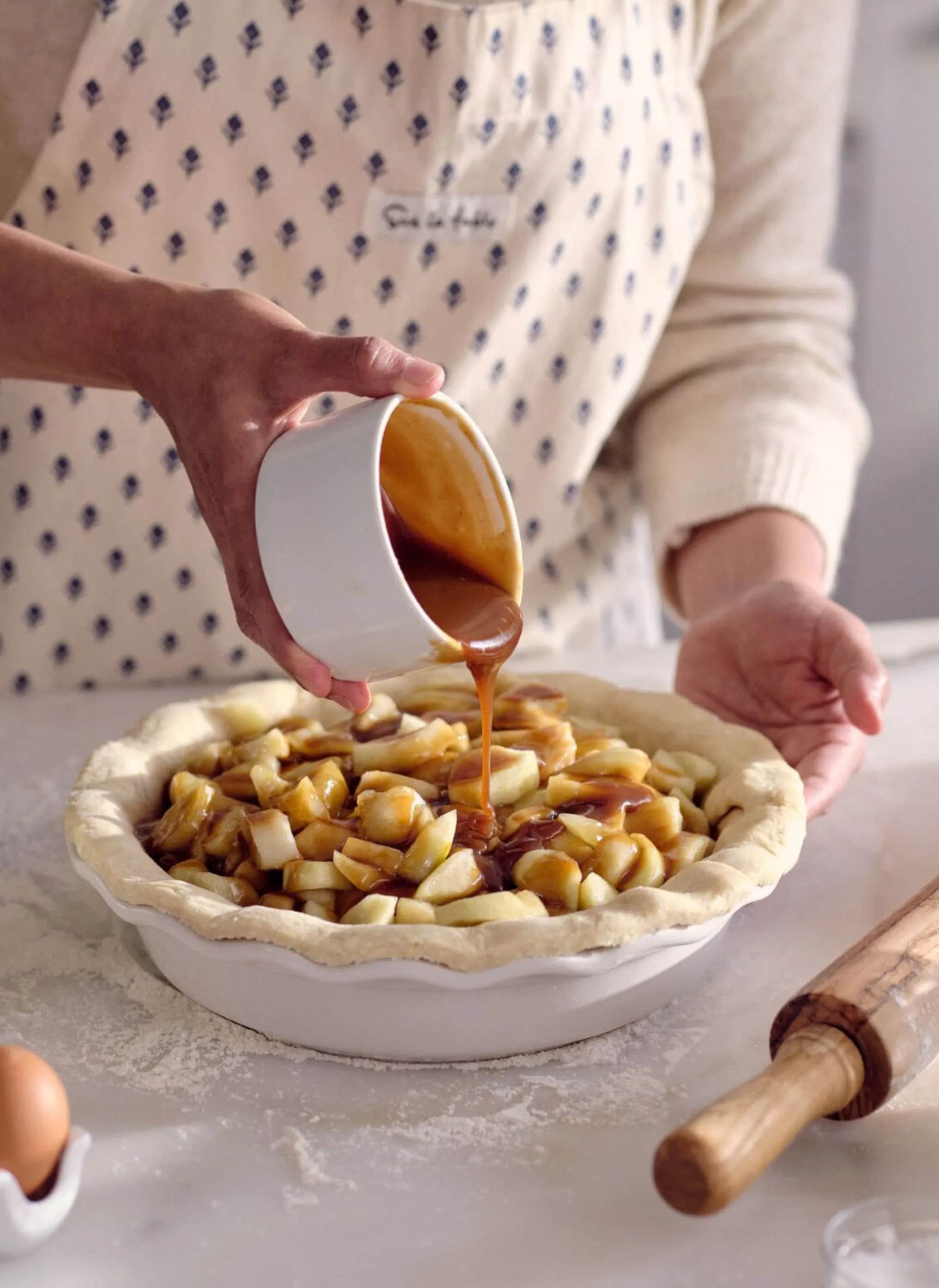 Person in apron pouring caramel over apple pie, with rolling pin nearby