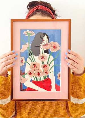 Person holding a framed colorful illustration of a woman with flowers and clouds, partially covering their face