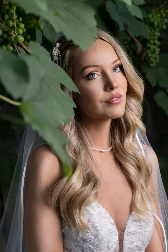 Bride in white gown with veil, posing among green leaves, looking thoughtful