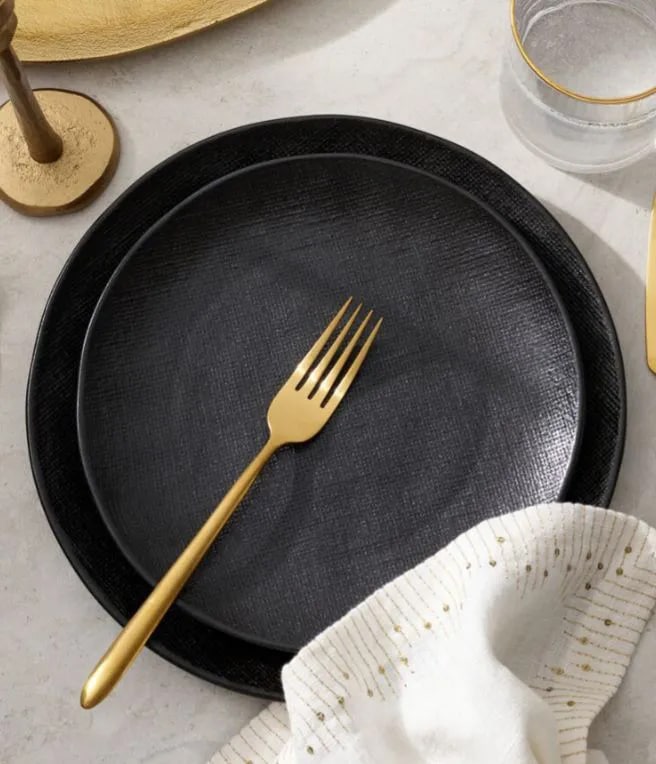 Black plate with a gold fork on a tablecloth, with a glass and utensils in the background