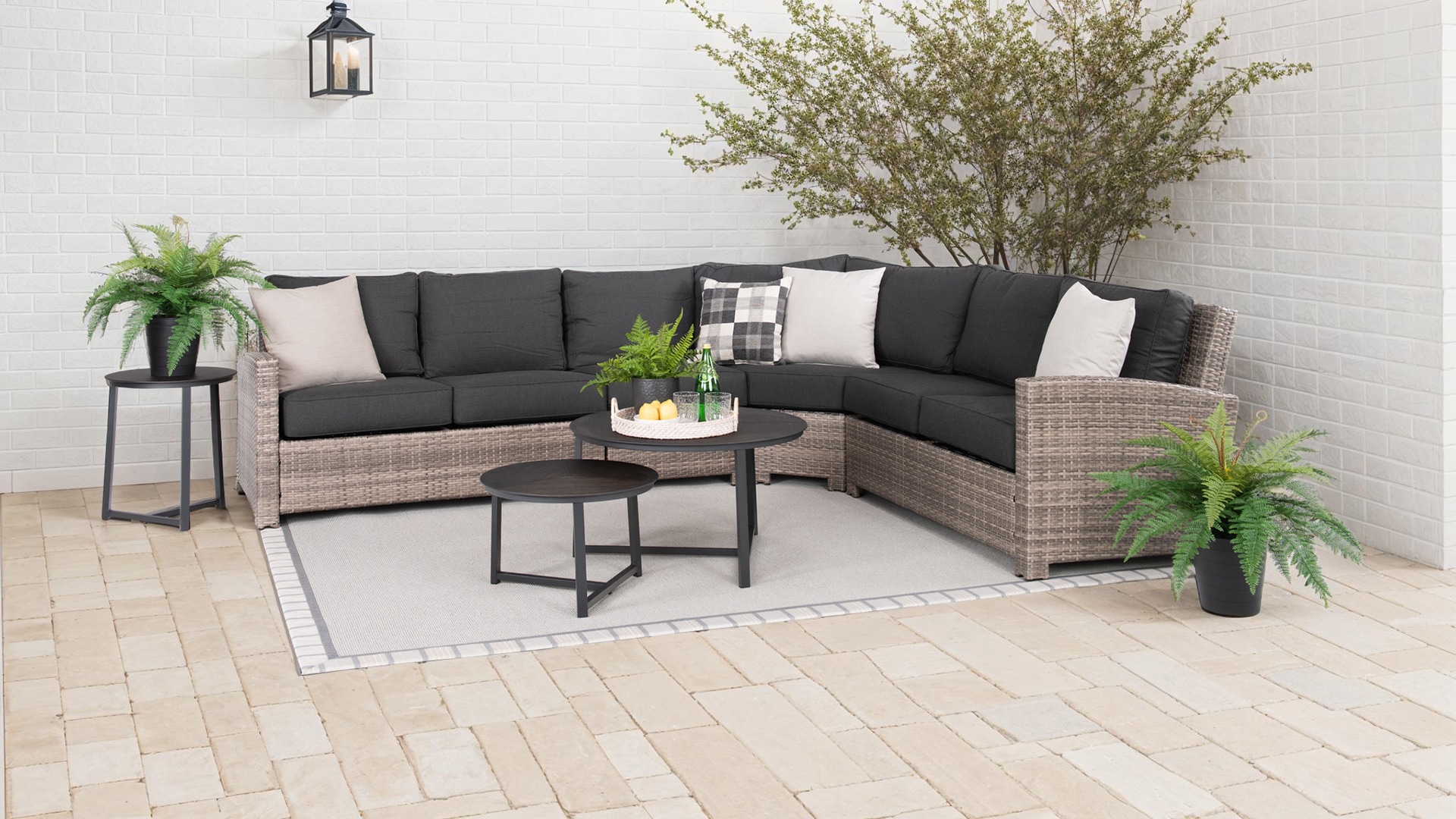 Outdoor patio setup with L-shaped sofa, two side tables, and a coffee table, decorated with plants