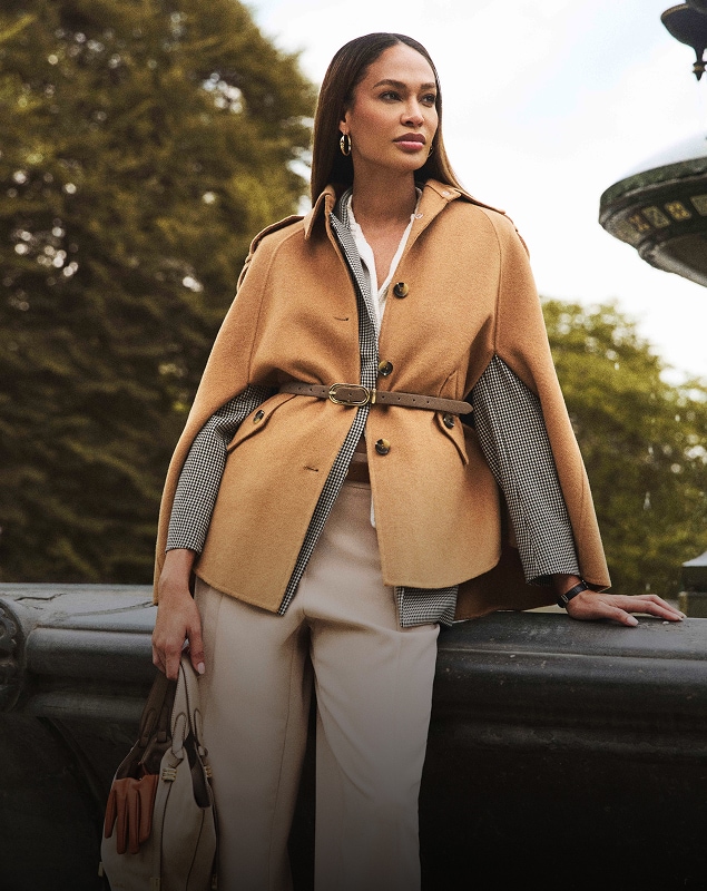 Woman in a stylish beige cape and trousers holding a bag, standing outdoors