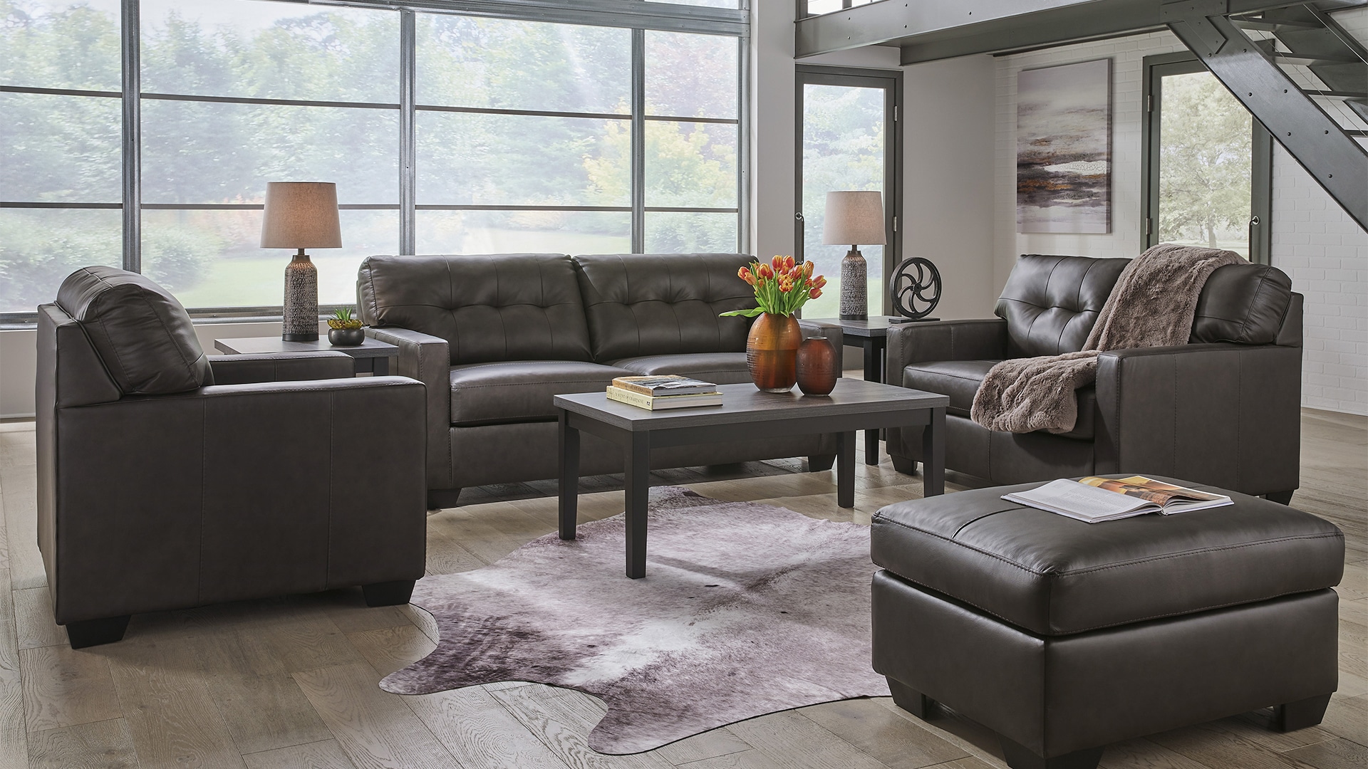Modern living room with a dark sofa set, armchair, ottoman, and a wooden coffee table, large windows in the background