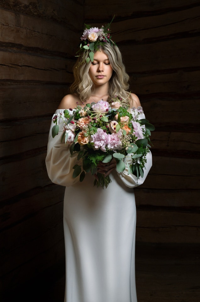 Woman in white dress with floral crown holding bouquet, standing against wooden background