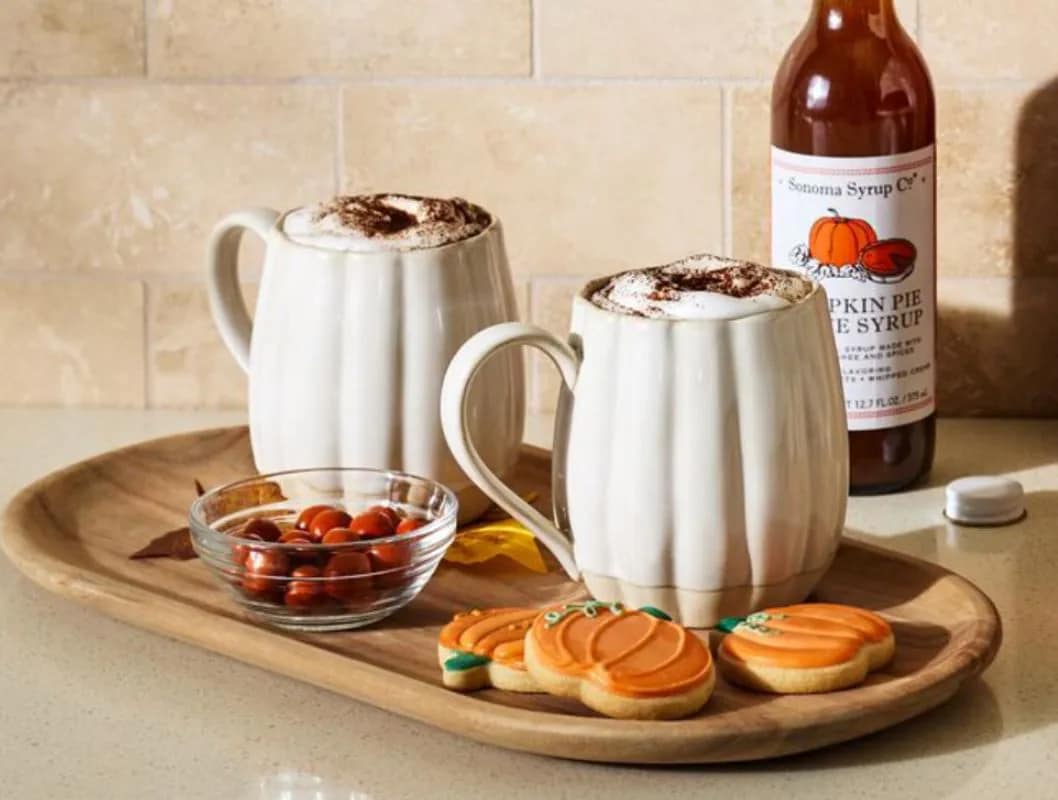 Two mugs of hot chocolate with whipped cream, cookies, almonds on a tray, and a bottle in the background