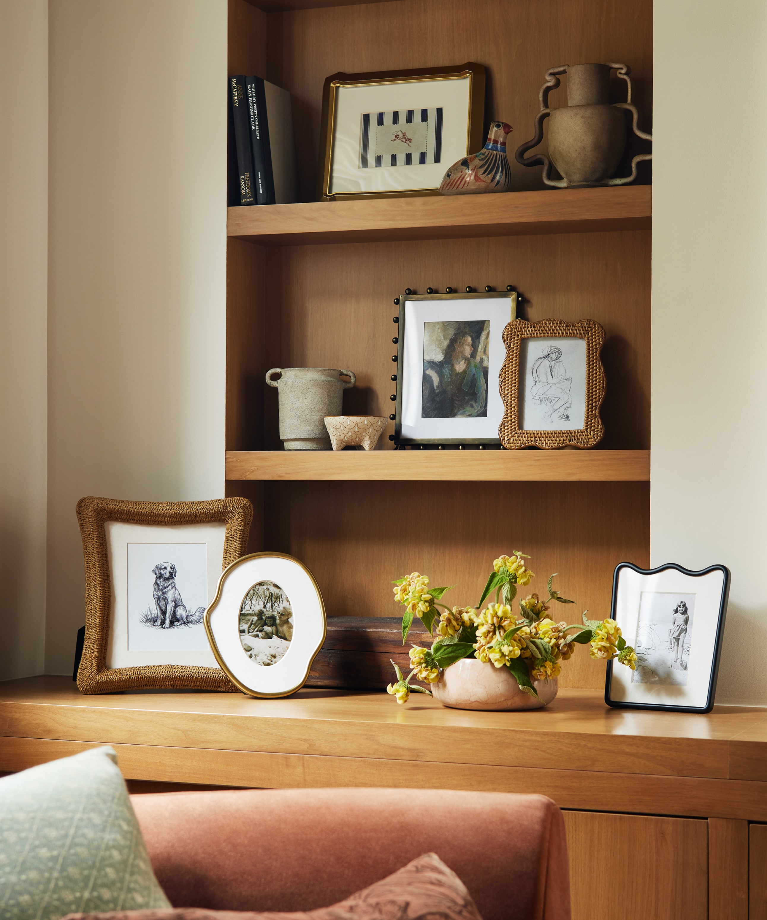 Cozy nook with a wooden shelf displaying framed photos, pottery, and books, over a cushioned bench