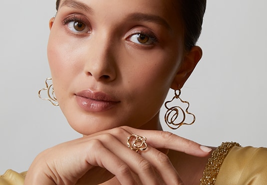 Woman with curly updo, showcasing intricate earrings and a ring, wearing a gold blouse