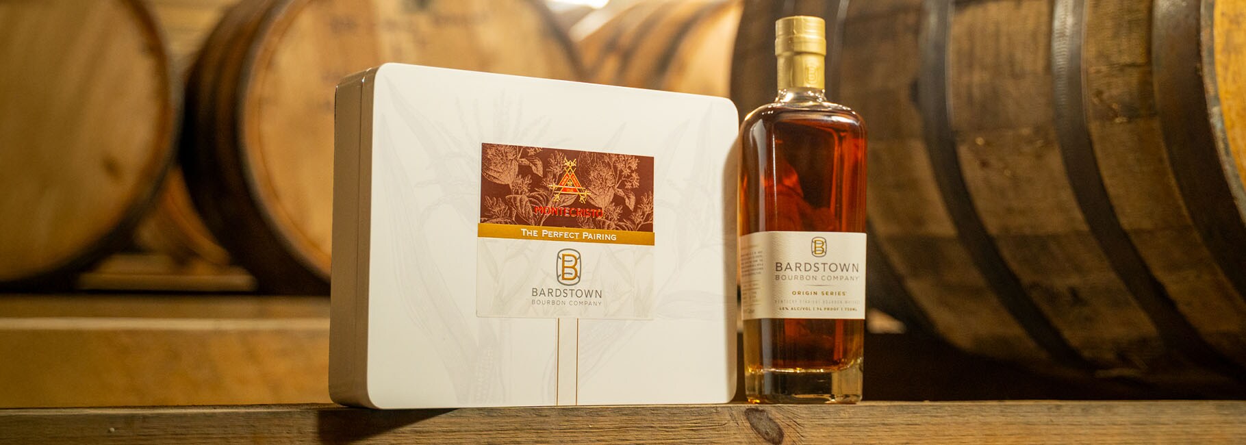 A bottle of Bardstown whiskey with a box of Bardstown cigars, on a wooden table, with barrels in the background