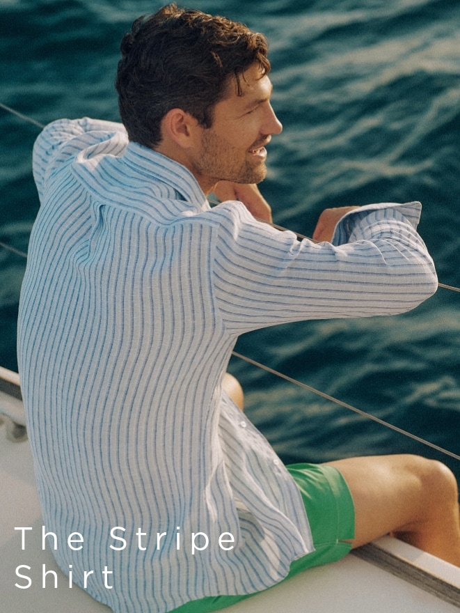 Man in a striped shirt and green shorts sitting on a boat, looking at the ocean