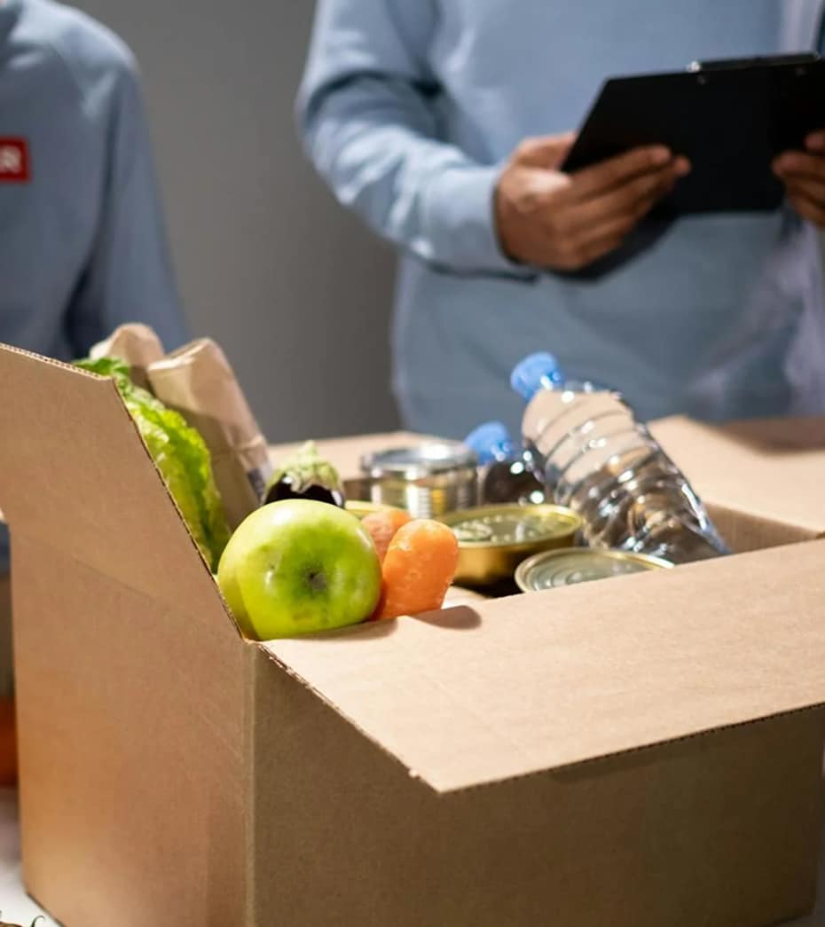 Open cardboard box with fruits, water bottles, and canned food, two people in background with clipboard