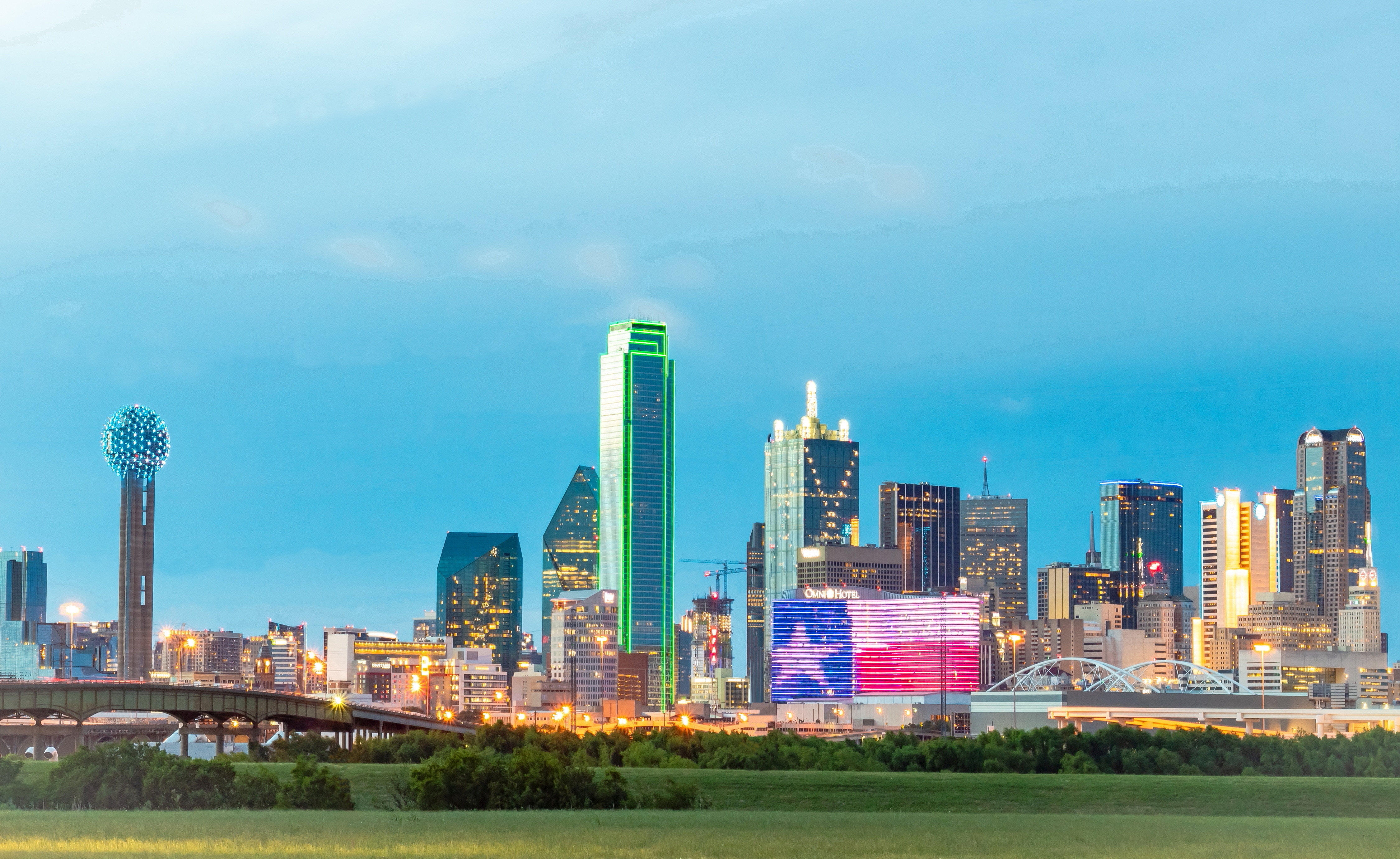 City skyline with tall buildings against blue sky, greenery in foreground