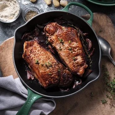 Two steaks cooking in a skillet with onions, garnished with herbs