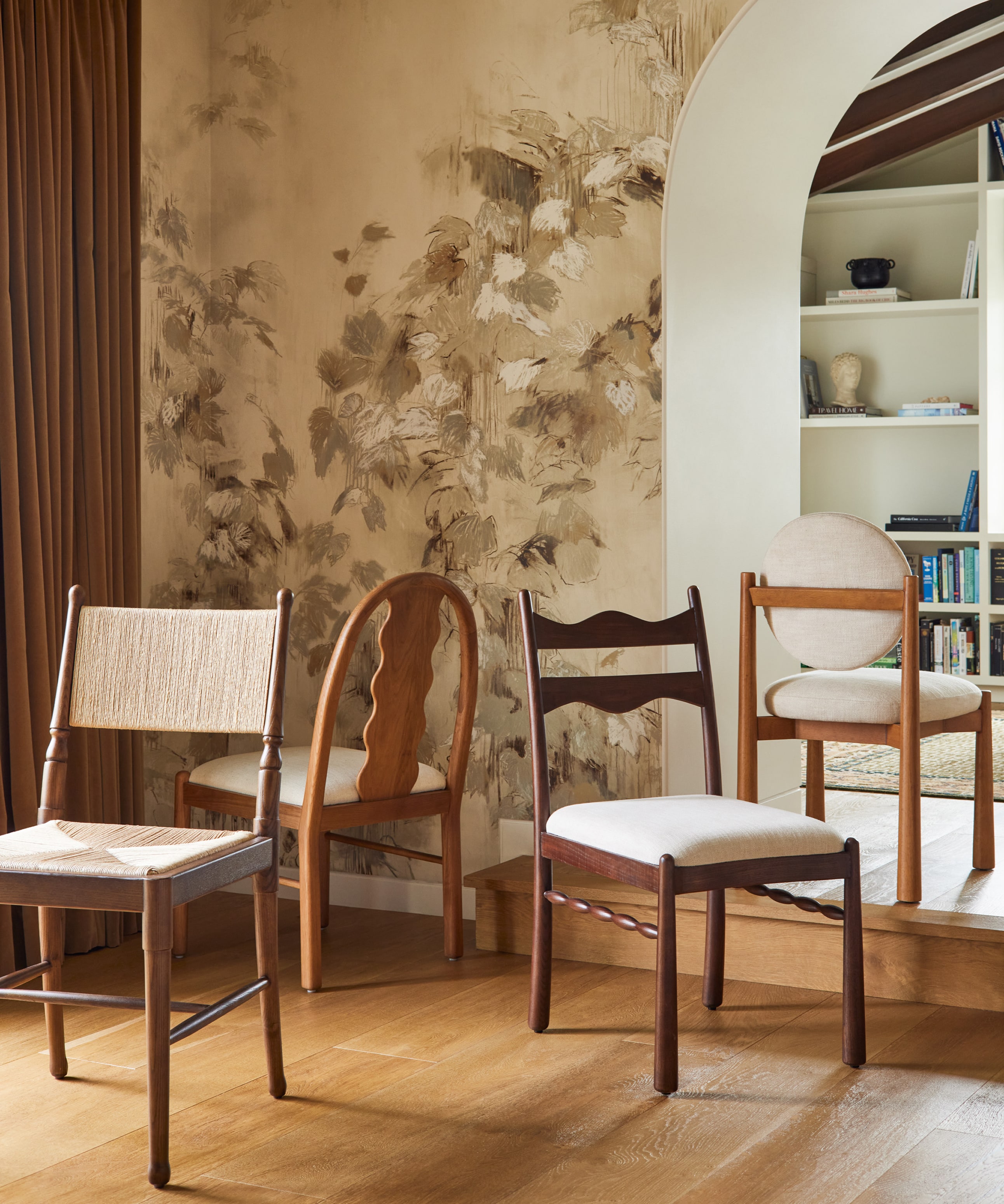 Four diverse chairs in a room with floral wall artwork and an archway leading to a bookshelf