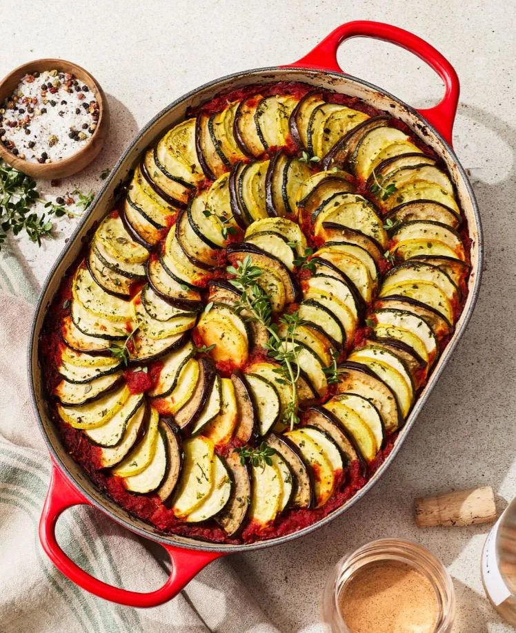A colorful ratatouille dish in a red pot, arranged in a spiral pattern of sliced zucchini and tomatoes