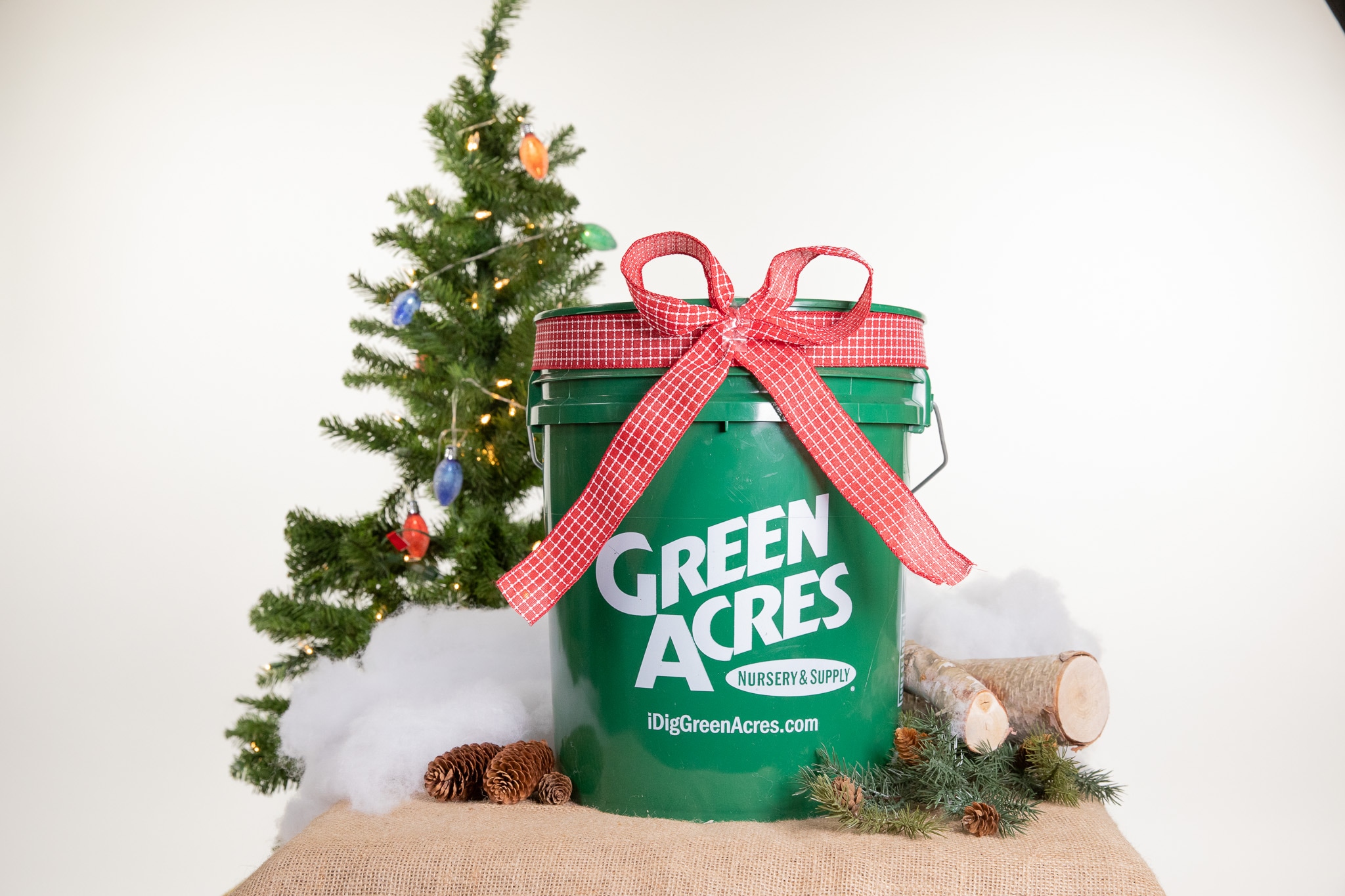 Green Acres-branded bucket with a red bow next to a Christmas tree