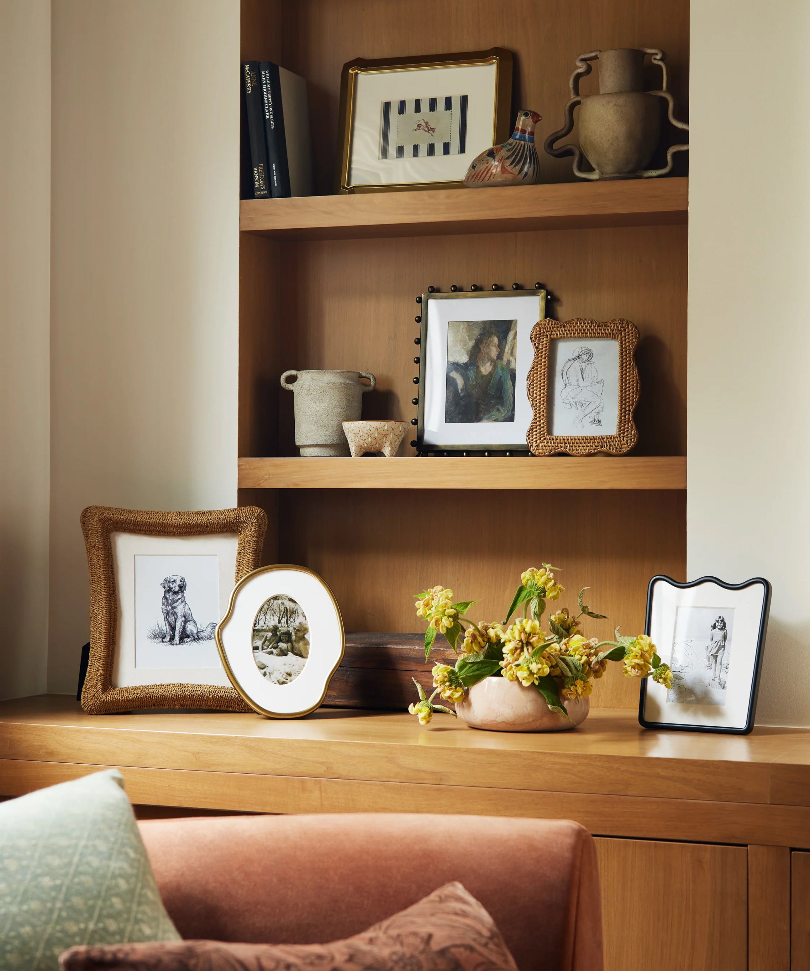 Warm living room corner with a wooden shelf displaying framed pictures, books, and pottery, adjacent to a velvet sofa