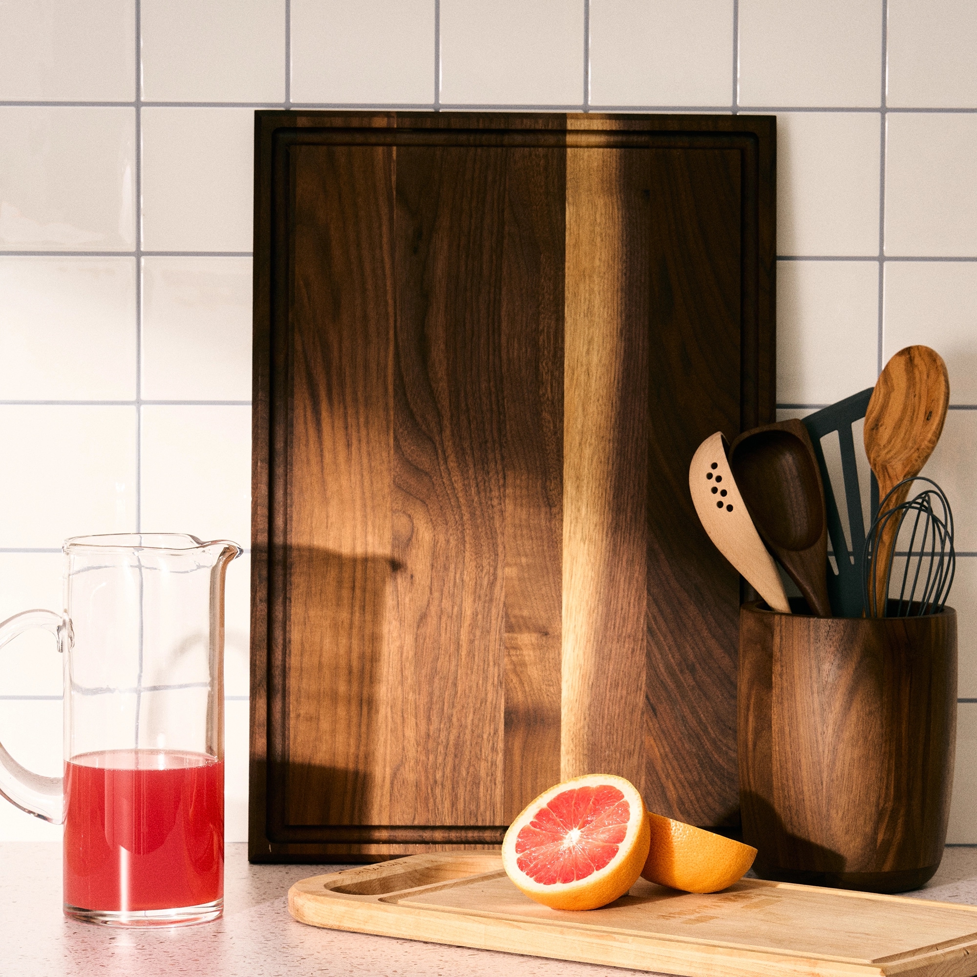 Wooden cutting board with kitchen utensils in a holder, a sliced grapefruit, and a glass of juice on a counter