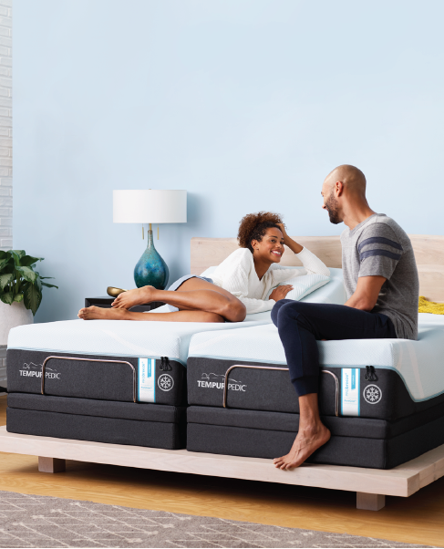 A couple smiling and talking on a mattress in a bright bedroom