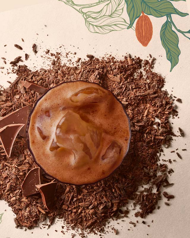 A cappuccino with a milk foam swirl surrounded by chocolate shavings and chocolate pieces on a decorated surface