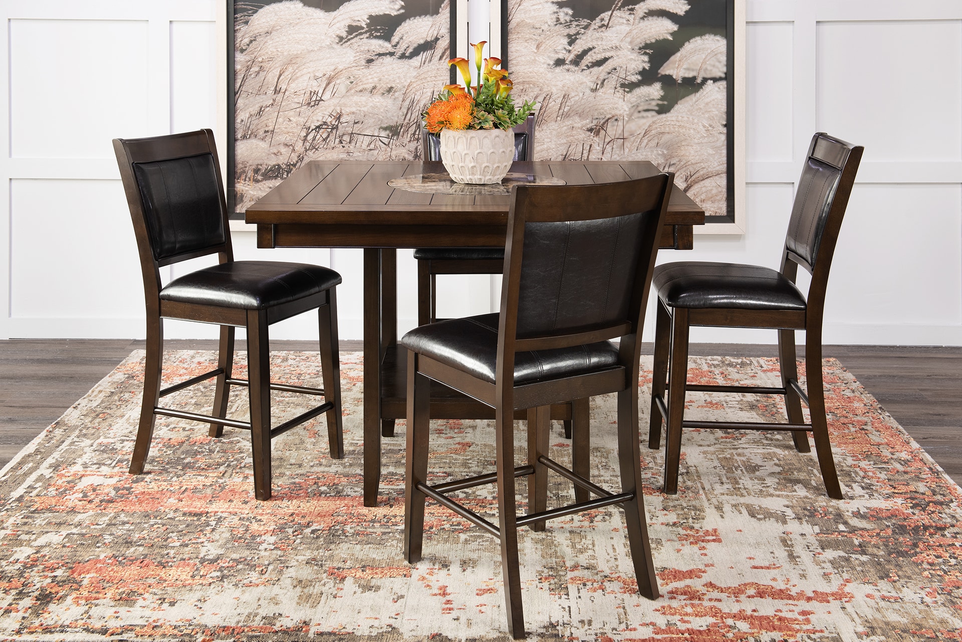 A dining table with four chairs on a patterned rug, positioned against a wall with art