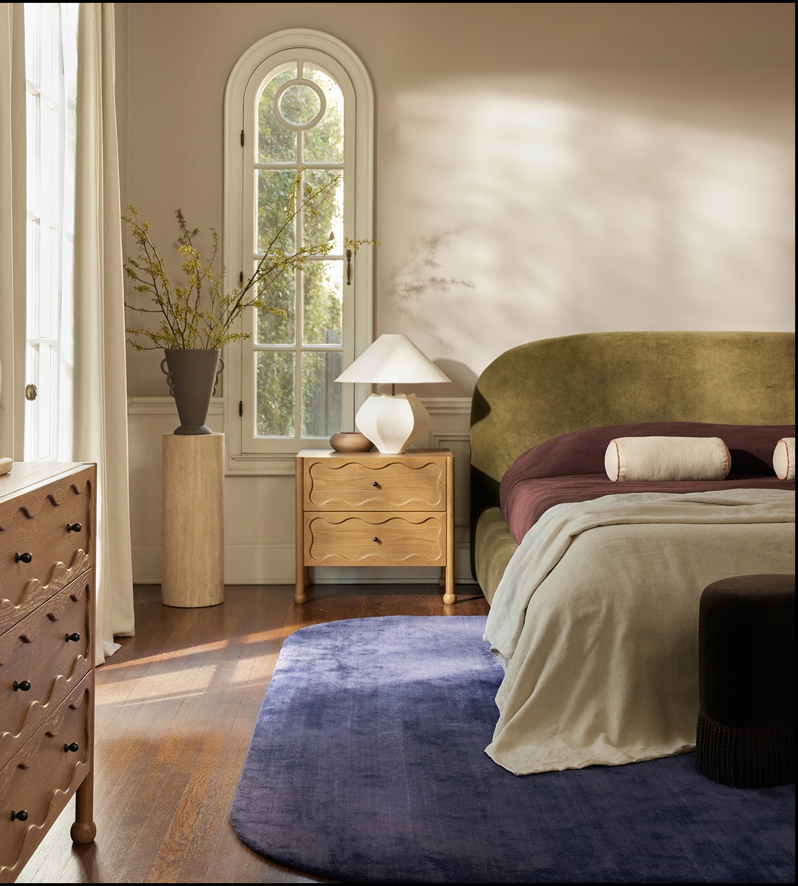 Elegant bedroom with a velvet bed, wood drawer, round lamp, and a blue rug under sunlight from a tall arched window