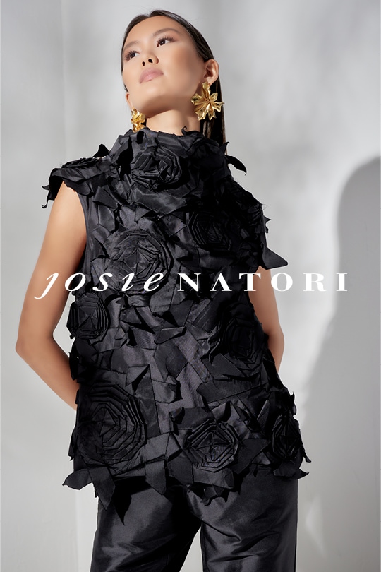 Woman in black ruffled outfit and gold earrings posing with Josie Natori logo