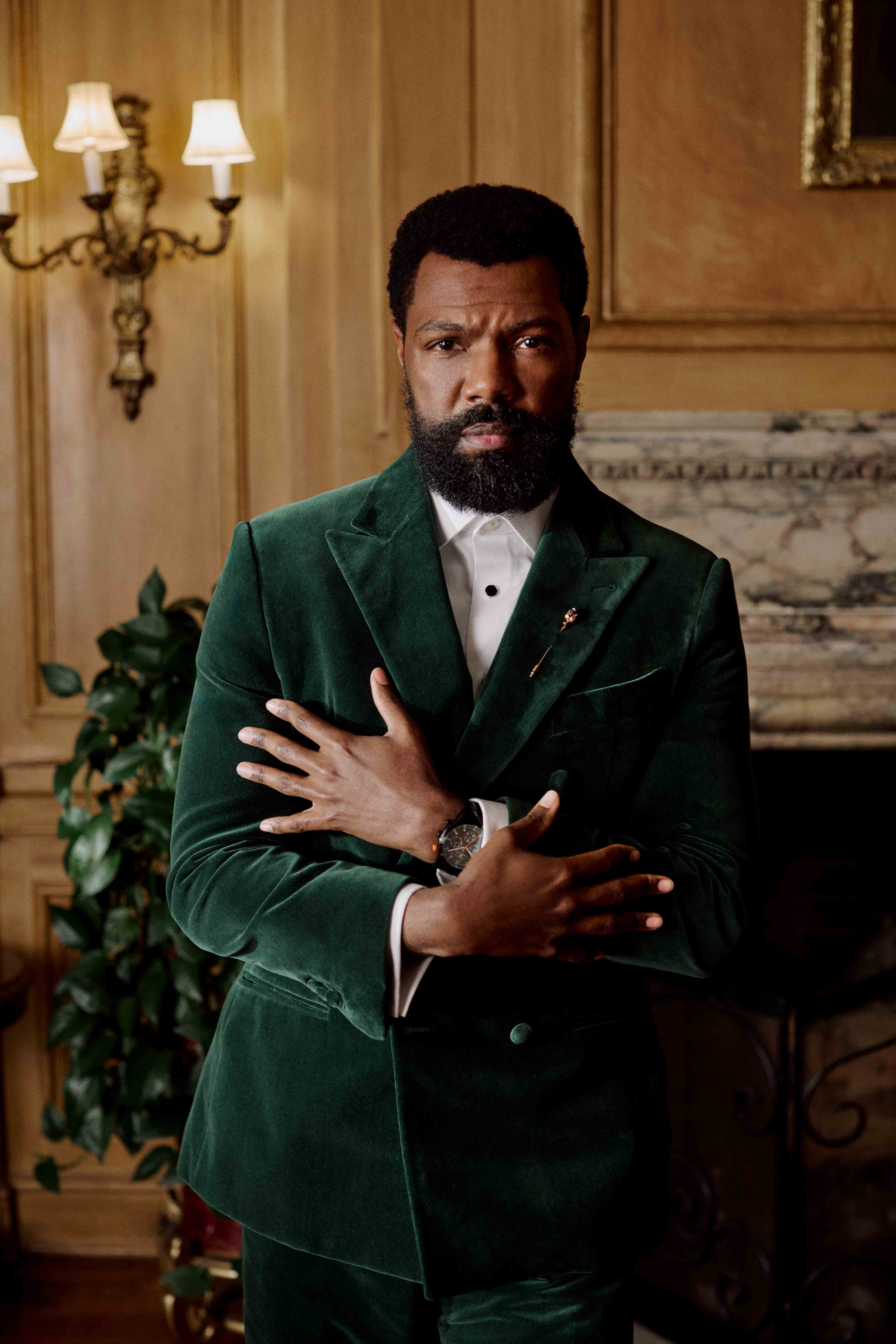 Man in a green velvet suit standing with arms crossed in an ornate room