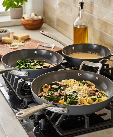 Three All-Clad nonstick skillets with helper handles on gas cooktop