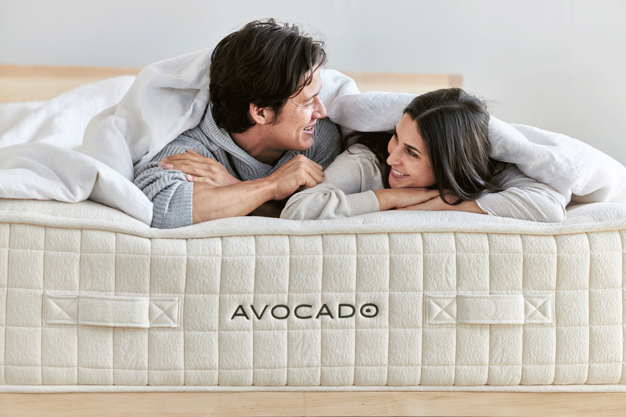 Couple smiling at each other in bed with "Avocado" branded mattress visible