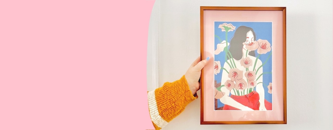 Person's hand reaching for framed artwork with a woman and cat, against a pink background