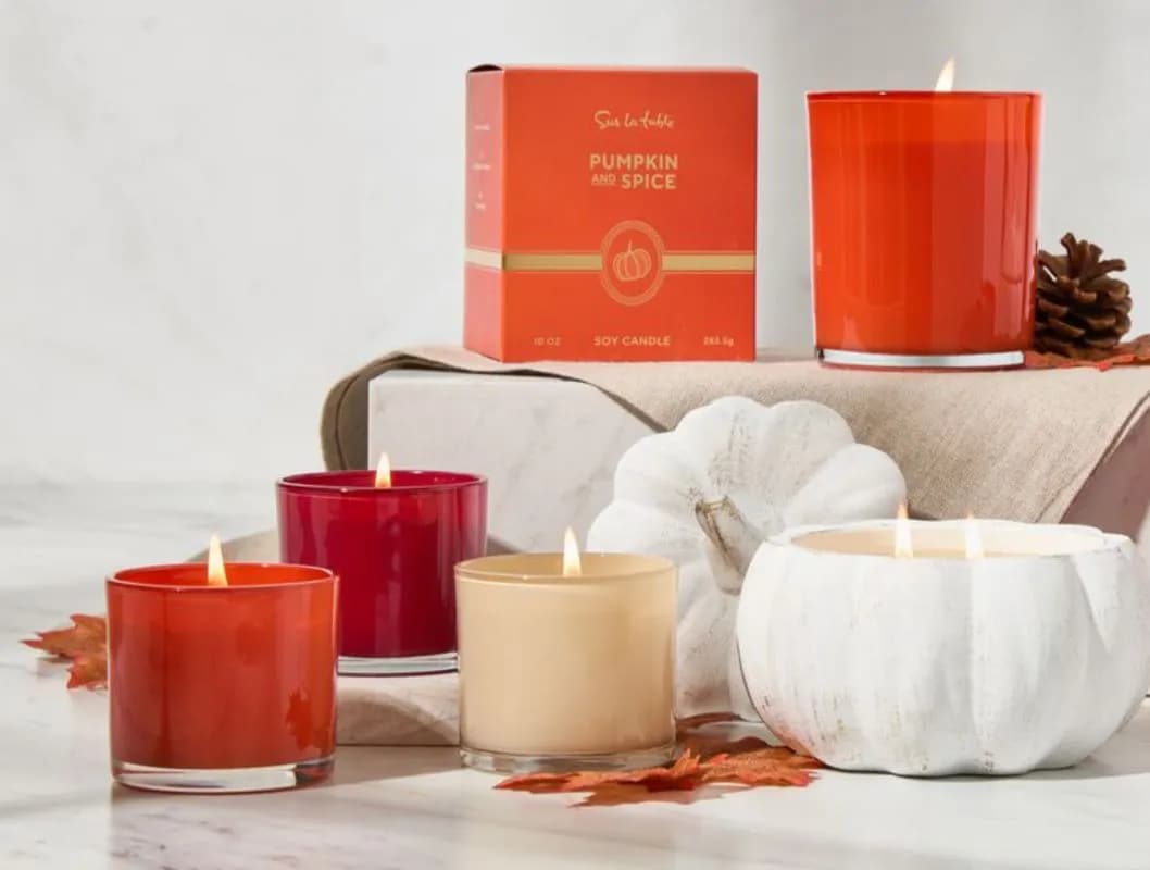 Autumn-themed display with scented candles in red and cream, white pumpkin, dried leaves, and decorative books