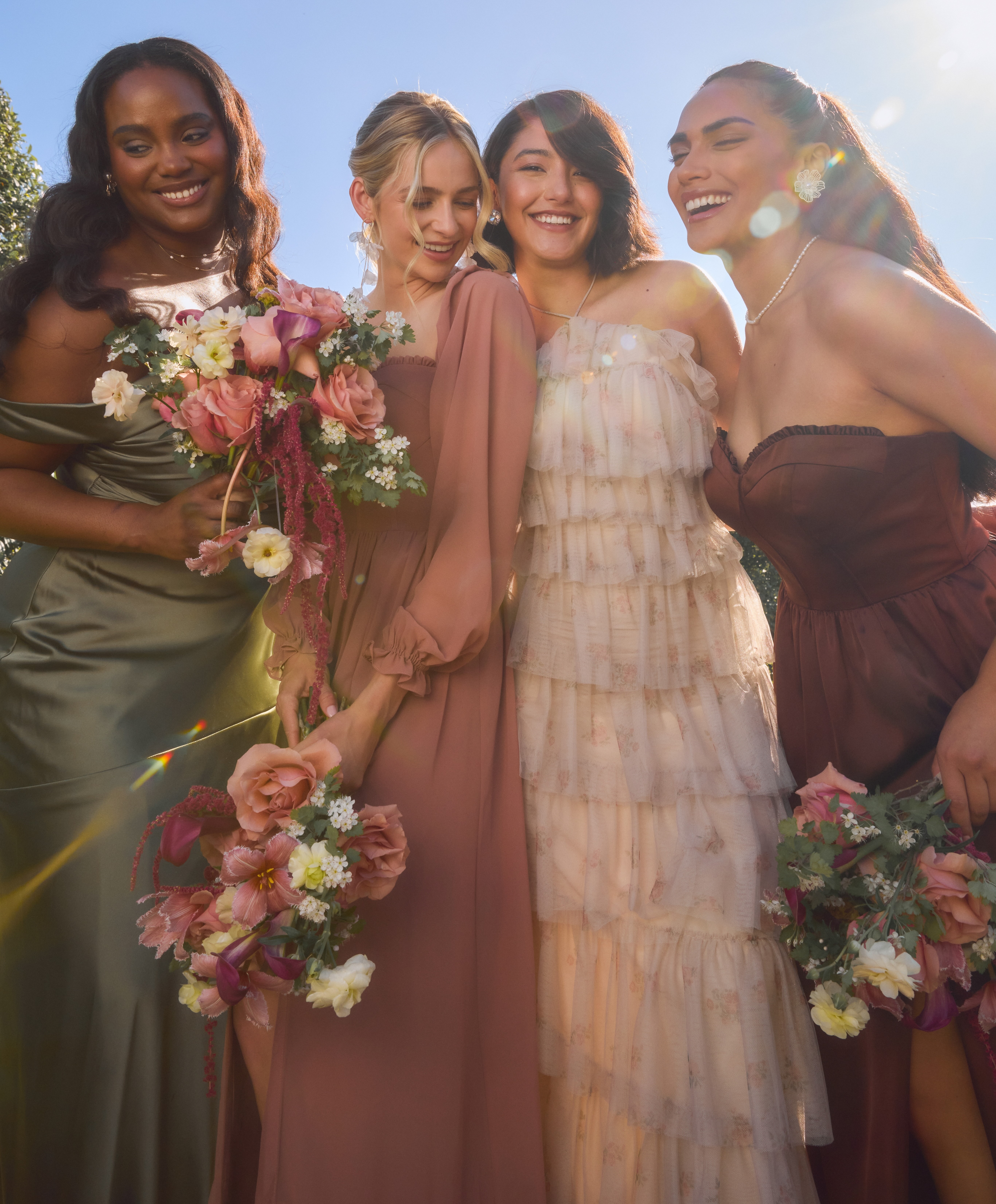 Four bridesmaids in pastel dresses holding bouquets, joyfully posing under sunlight