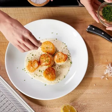 A person seasoning seared scallops on a white plate