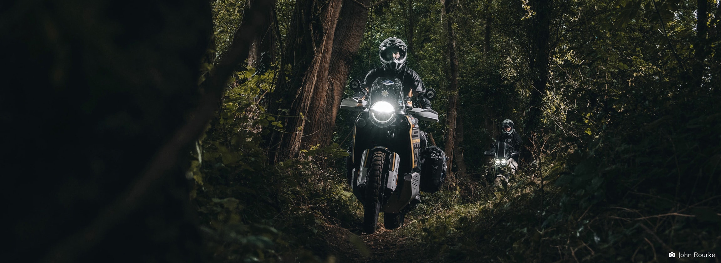 Rider on a motorcycle with headlights on, driving through a shadowy forest