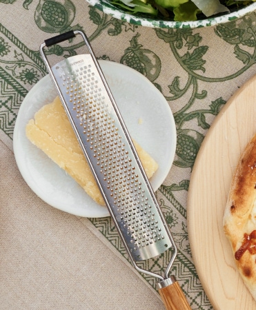 Metal grater resting on a lemon slice and plate, near a wooden serving board with pizza