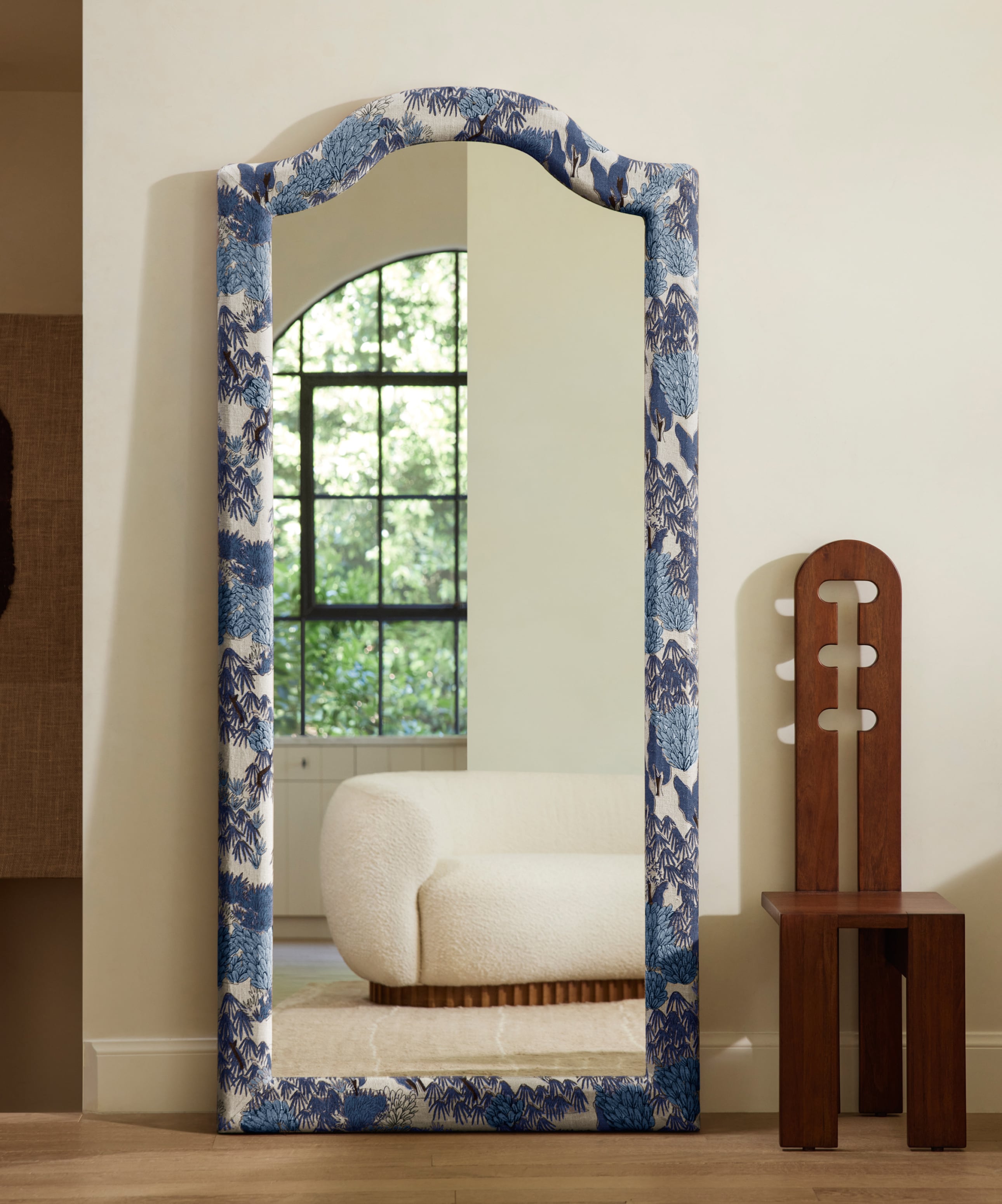 Tall arch-top mirror with blue floral frame in a room reflecting a window and chair