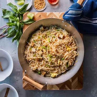 Bowl of stir-fried noodles with vegetables, chopsticks on side, ingredients around, on a table