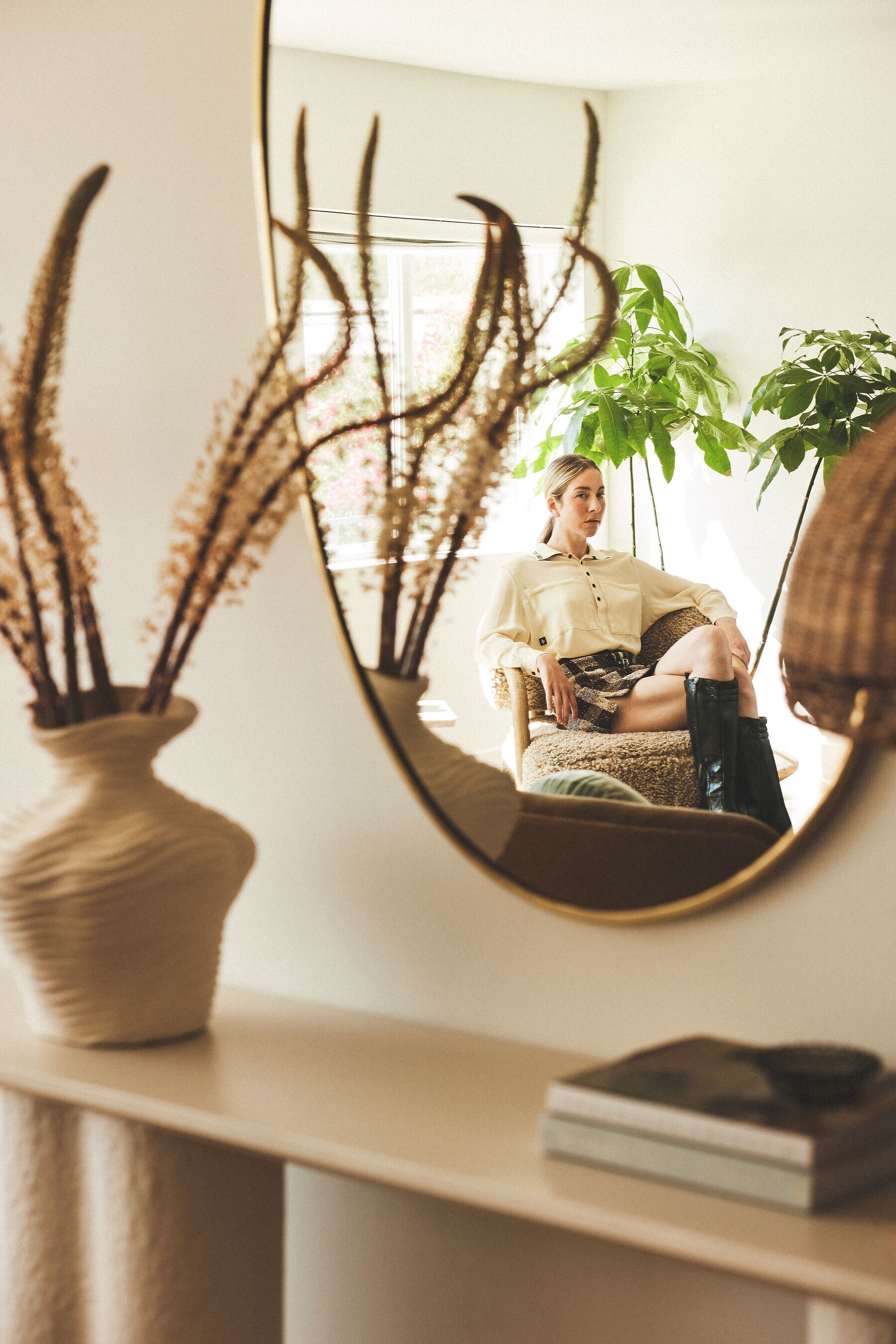 Woman reflected in a round mirror, sitting with a book, surrounded by plants and dried flora