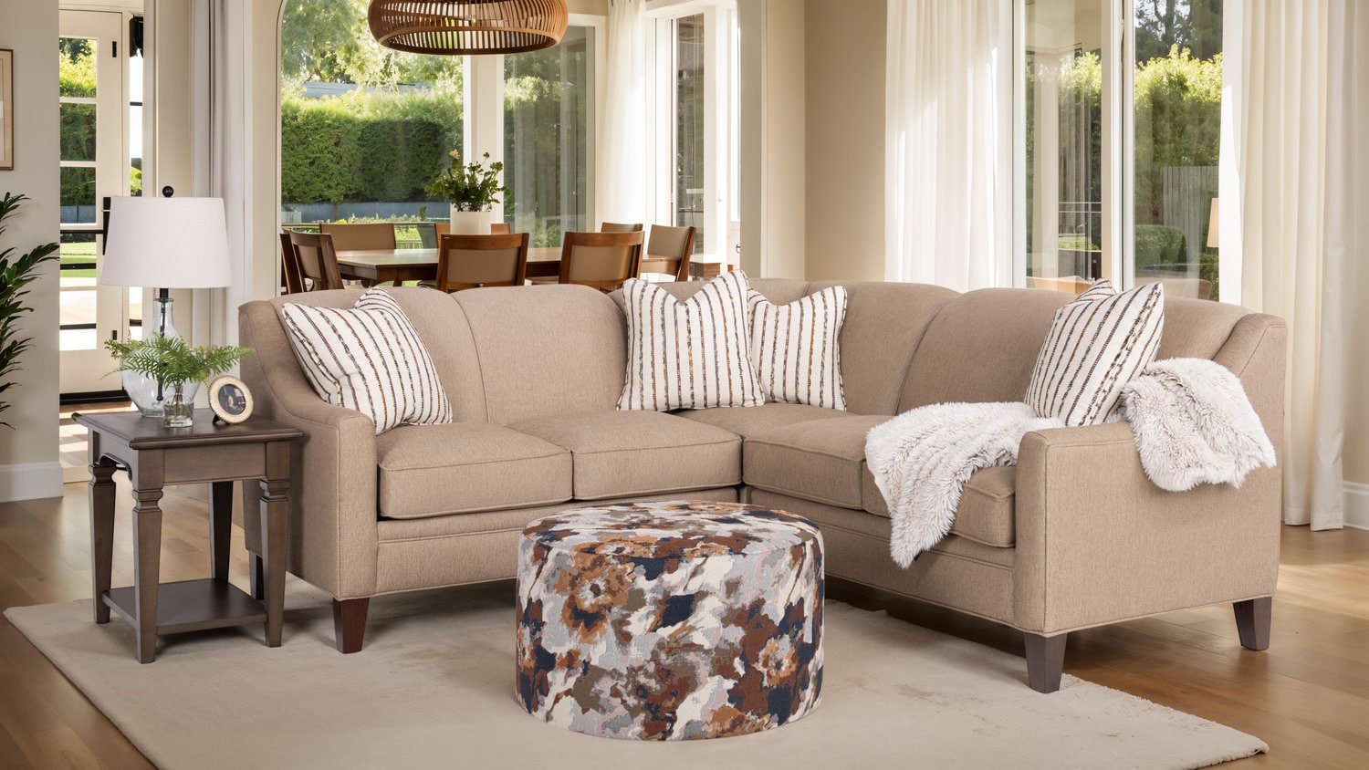 Elegant living room with a beige sofa, striped pillows, round floral ottoman, side table, and large windows