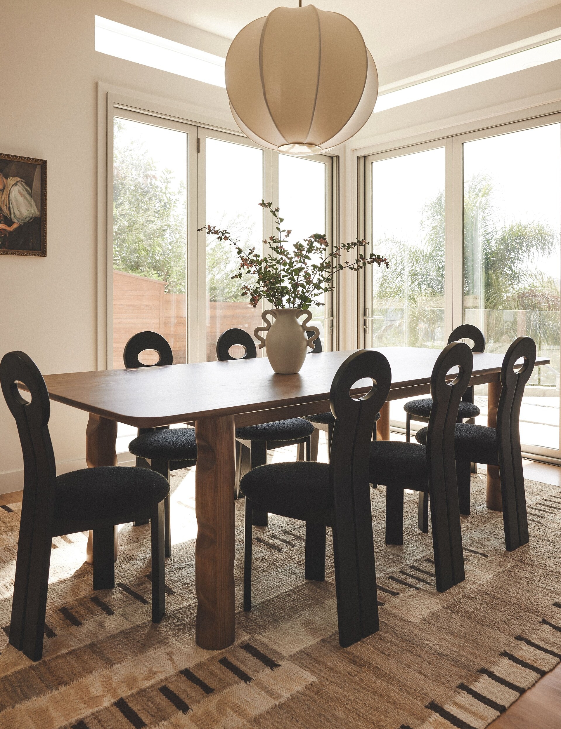 A bright dining room with a wooden table, black chairs, large windows, and a decorative lamp