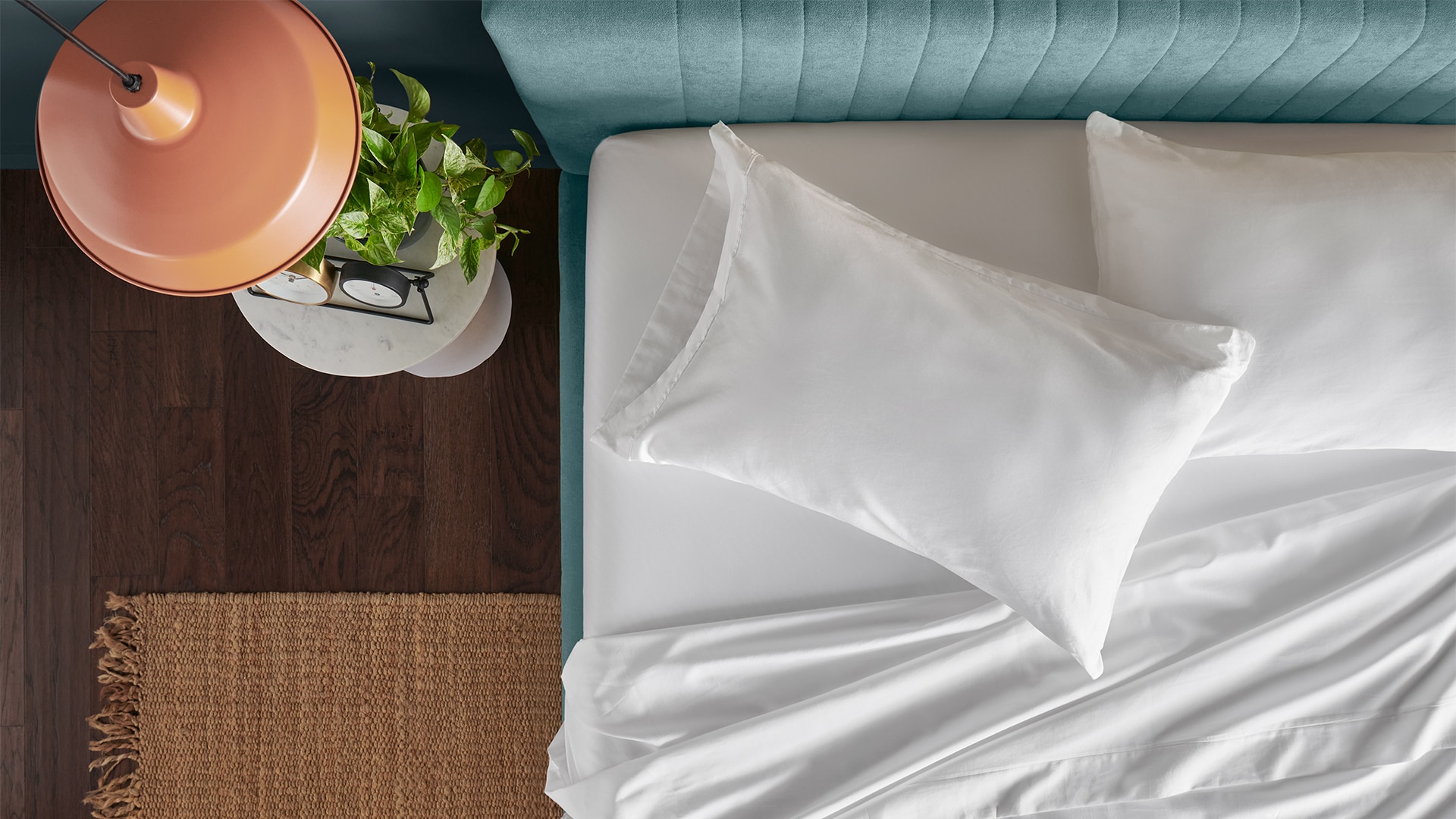 Top-down view of a neatly made bed with teal headboard, white bedding, and a lamp and plant on a bedside table