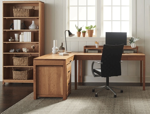 A well-lit home office with a wooden desk, chair, and bookshelf, decorated with plants and simple decor